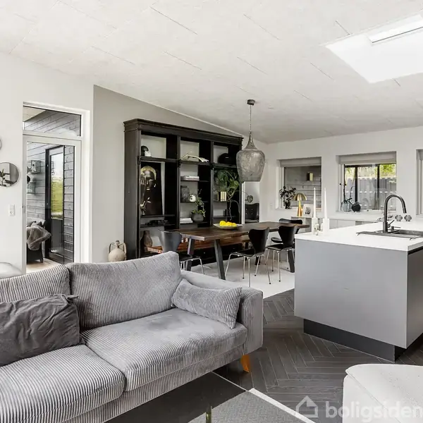 Grå sofa står i moderne stue-køkken-alrum. Køkkenø med integreret vask, spisebord med stole, store vinduer, og adgang til terrasse. Indretningen er lys og minimalistisk.