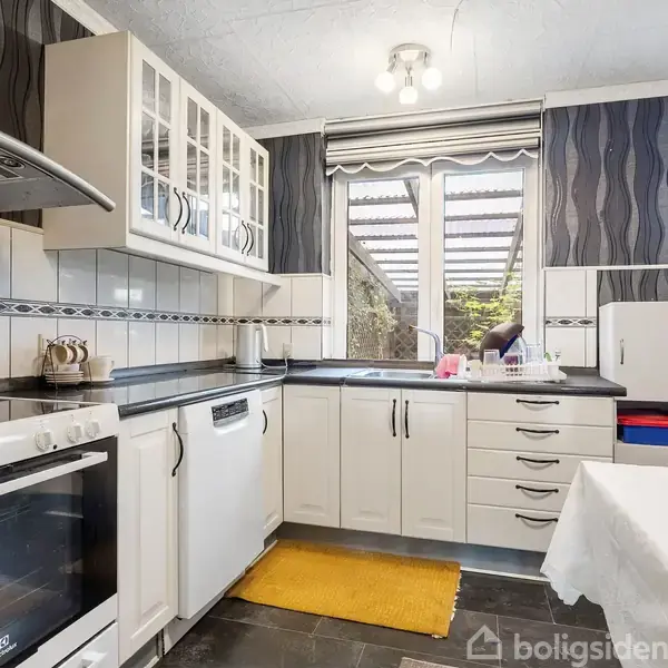 Køkken med hvide skabe og skuffer, sort bordplade og komfur. En gul måtte ligger på gulvet. Store vinduer giver udsigt til en overdækket terrasse. Bord med hvid dug i forgrunden.