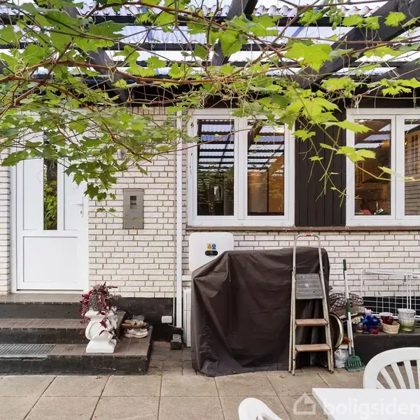 Dør og vinduer i et hvidt murstenshus. Foran døren findes blomster i potter, og en grill dækket af et sort betræk står tæt på. Plastikhavemøbler står på fliser.