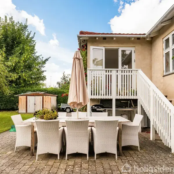 Hus med rødt tag og terrasse, omgivet af stor, grøn have med træer. Naboer, villaer og veje ses i baggrunden.