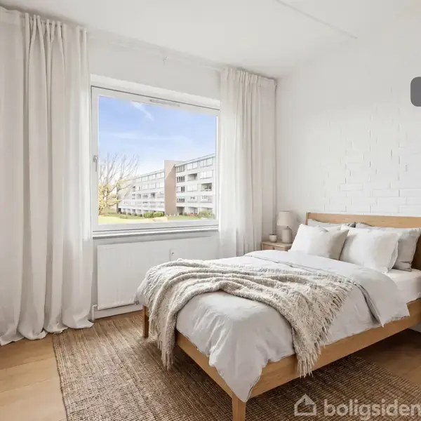 Soveværelse med en dobbeltseng dækket af tæpper og puder. Sengen står ved siden af et natbord med en lampe og planter. Store vinduer med gardiner viser bygninger udenfor. Tekst: "Visualisering".