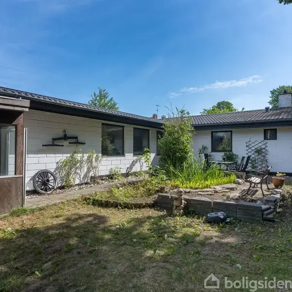 En hvid villa med fladt tag står i en have. En stenbænk og planter omkranser et lille anlæg. Solrig dag med en blå himmel.