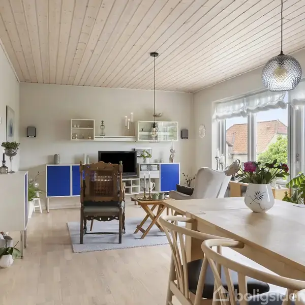 Stuen har et lyst og luftigt interiør. Et langt spisebord med træstole står i forgrunden. Store vinduer til højre giver udsigt til en frodig have.