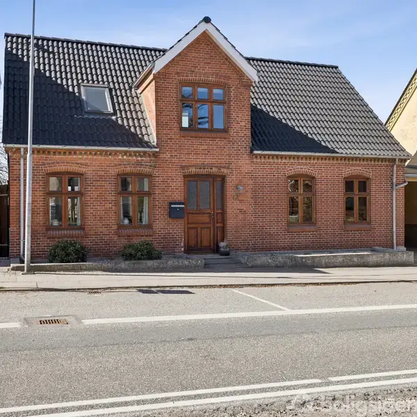 Rødt murstenshus med sorte tegltag, tre frontvinduer og centralt dørparti. Det står på en fortovsgade med tilstødende bygninger. Der er ingen synlig tekst.
