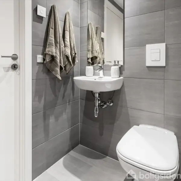 Et lille badeværelse med grå fliser på væggene. En vask med sæbebeholder og to tandkrus er monteret under et spejl. Ved siden af hænger håndklæder, og der er et toilet.