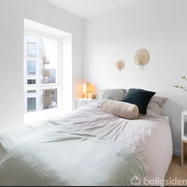 Seng med puder står i et lyst soveværelse ved et vindue. To væglamper hænger over sengen, og en natbord med dekorationer står til højre.
