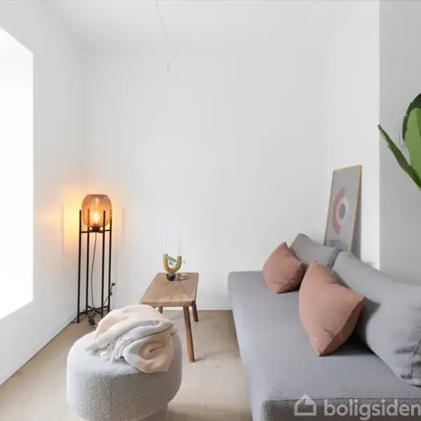 En grå sofa med puder står i et lyst værelse. Ved siden af er en plante og et lille bord med en drink. En skammel og lampe står tæt ved vinduet.