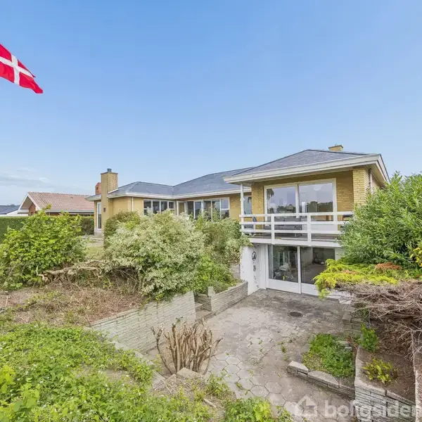 Et hus med gulstensfacade står midt i en frodig have. En flagstang med Dannebrog svajer i vinden. Huset har en balkon med udsigt.