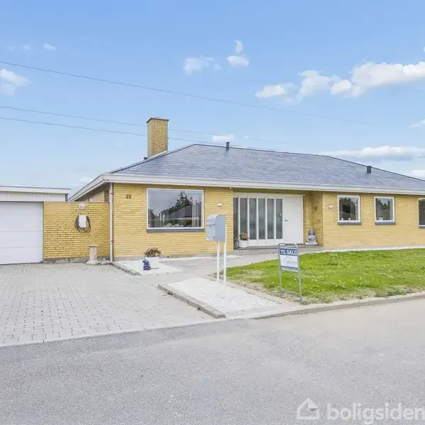 En gul murstensvilla med fladt tag og store vinduer står langs en rolig vej. Foran huset er en indkørsel, garage, og en græsplæne med skiltet "TIL SALG."