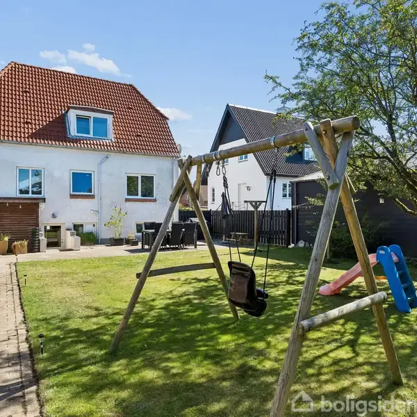 Et legehus står i græsplænen med en rutsjebane og gynger. I baggrunden ses et hvidt hus med rødt tegltag og flisebelagt terrasse, omgivet af en have.