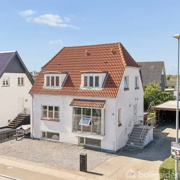 Et hvidt hus med rødt tegltag står langs en vej. Der er trapper op til hovedindgangen. Omkringliggende huse og parkeringsplads ses i baggrunden.