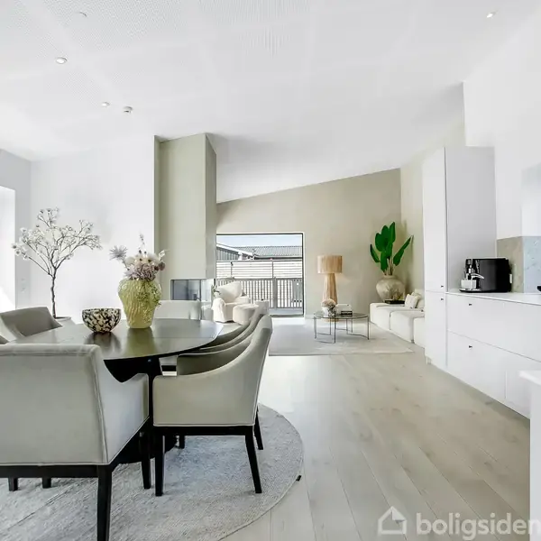Et moderne spisestue-sæt står på et lyst trægulv, indrammet af store vinduer. I baggrunden ses en sofagruppe og dekorationer, mens køkkenområdet har hvide elementer og en sort vandhane.