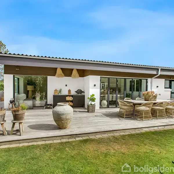 En moderne bungalow med fladt tag står på en træterrasse, hvor der er havemøbler og planter. Terrassen er omgivet af grønne buske og en græsplæne under en klar blå himmel.