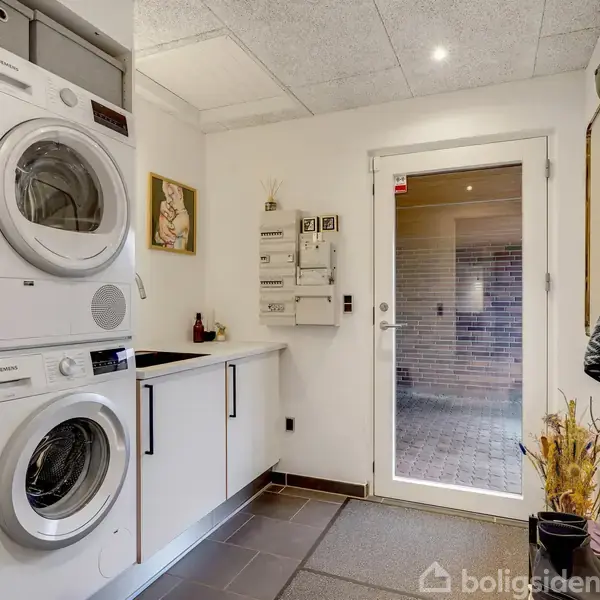 En vaskemaskine og tørretumbler står stablet i et bryggers. En dør fører ud til en flisebelagt gårdhave. Jakker hænger på væggen, og der er skabe samt en hylde.