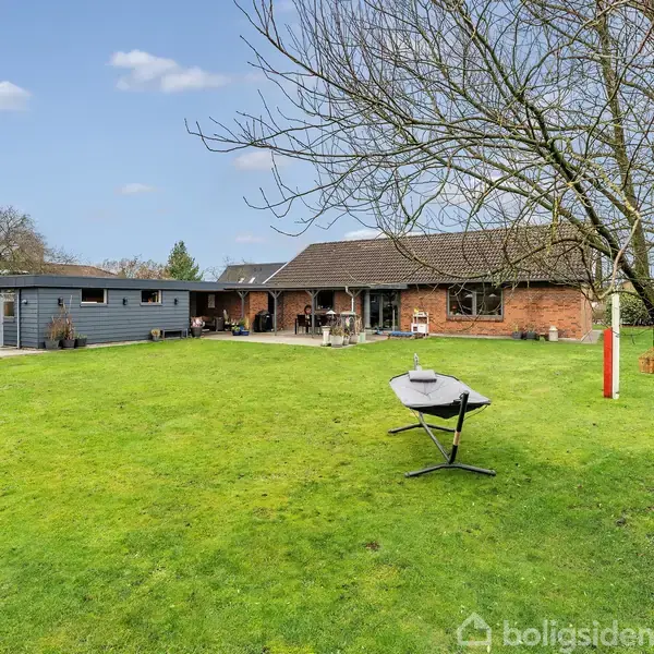 Græsplæne med et strygebræt i forgrunden, omgivet af et stort træ. I baggrunden ses et rødt murstenshus og en grå bygning under en blå, skyet himmel.