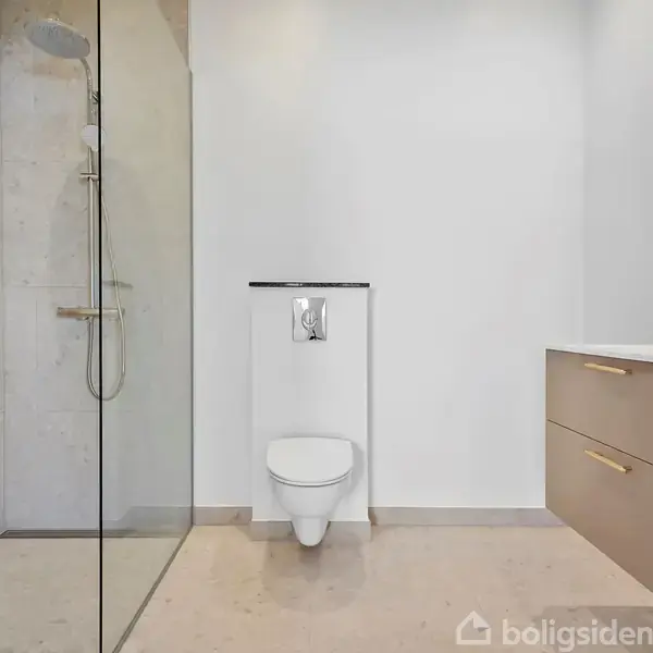 Et moderne badeværelse med en bruser til venstre, et væghængt toilet i midten, og en vask med et spejl til højre. Simpelt design med lyse fliser.