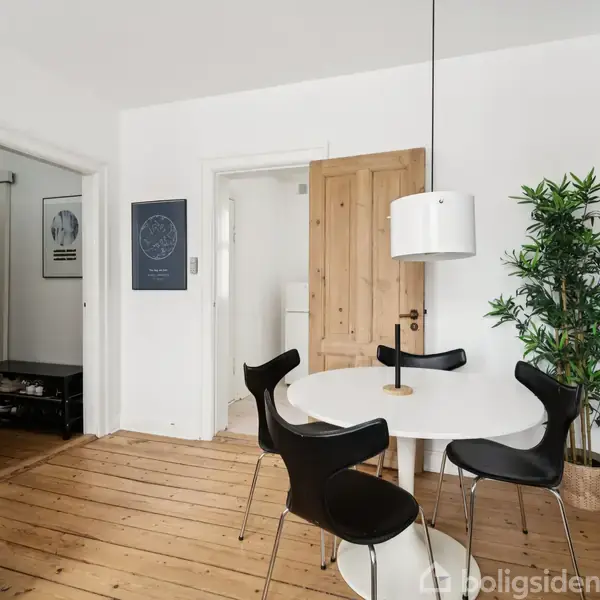 Et rundt hvidt spisebord med fire sorte stole står i et lyse rum med trægulv. En hængende lampe og en grøn plante tilføjer detaljer til indretningen.