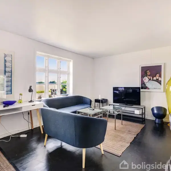 En stue med en blå sofa og et fladskærms-TV, placeret på et tæppe i et lyst rum med hvidmalede vægge og store vinduer. Et gult vase står i hjørnet.