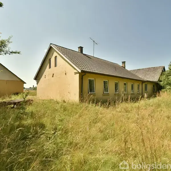 Tre landbrugsbygninger står i en hesteskoformet formation på en åben mark, omgivet af skov og græsarealer. En smal vej fører ind til komplekset fra højre.