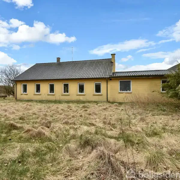 En gulstensbygning med fem vinduer står stille i et åbent område omgivet af vildtvoksende græs og spredte buske, under en blå himmel med få skyer.