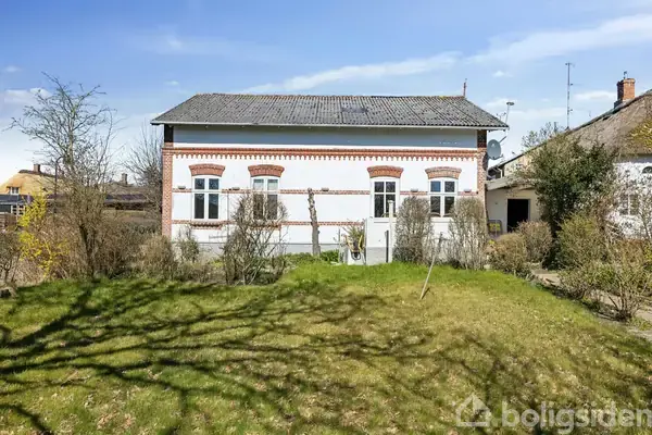 Hus med hvid facade og røde murstensdetaljer, står i en græsbelagt have omgivet af buske og træer; en blå himmel danner baggrunden.