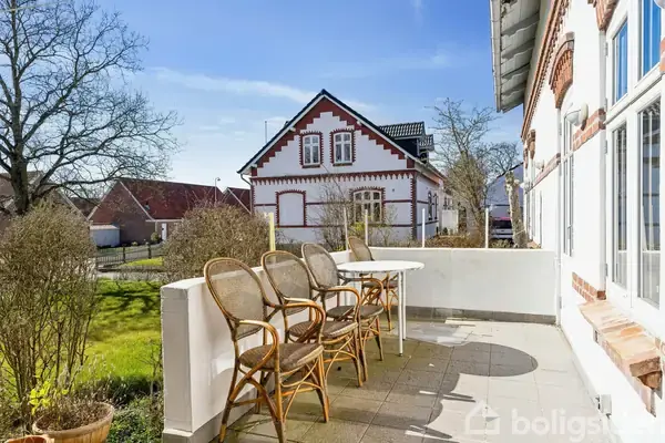 Terrassen har stole opstillet omkring et rundt bord. Den vender ud mod en have med et gammelt hus i baggrunden. Det er en solrig dag med blå himmel.