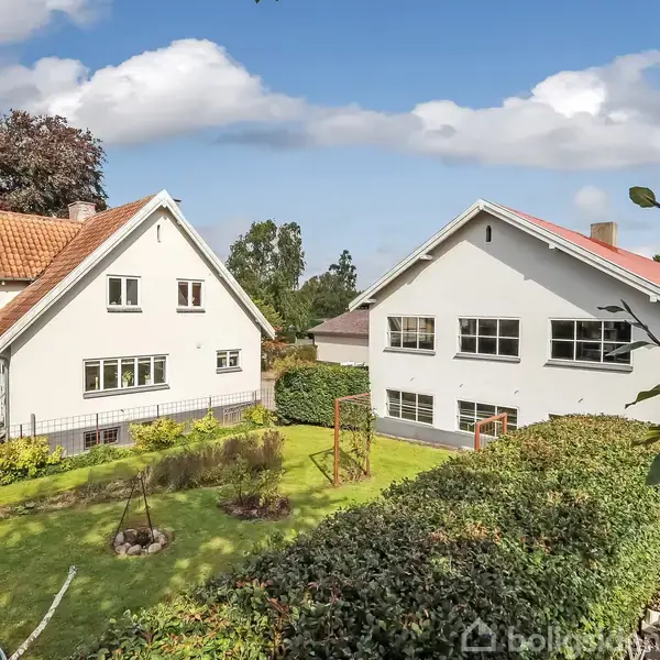 To huse med røde tegltage står i en frodig have med træer og buske under en blå himmel med skyer. Det ene hus har et bredt vindue foran.