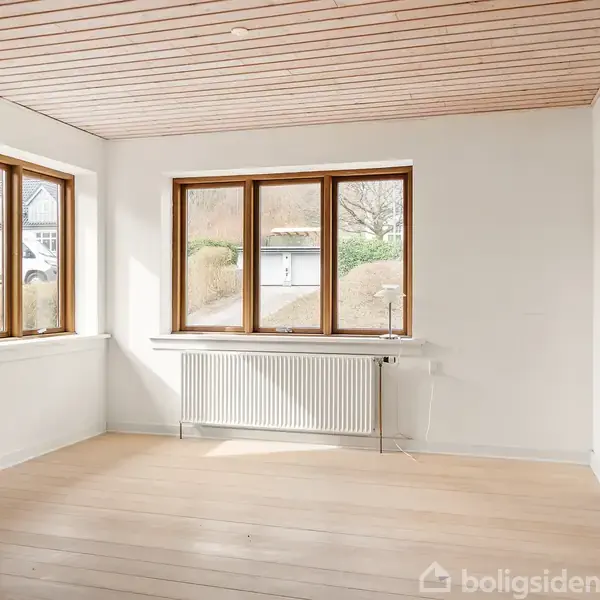 Et tomt rum med lyse trægulve og hvidmalede vægge, hvor sollys strømmer ind gennem store vinduer med trærammer. Væggene er hvide, og der er en radiator under vinduerne.