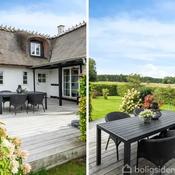 Traditionelt stråtækt hus med moderne møbler på en terrasse. Sort spisebord med stole står i fokus, omgivet af frodig have og åbne marker under en blå himmel.