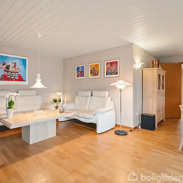 Hvid sofa og sofabord med blomsteraktion i lys stue; vægkunst og træspisebord i baggrund. Lamper hænger fra loftet, giver varm belysning. Indbydende rum med træparketgulv.