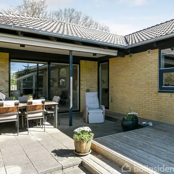 Terrasse med spisebord og stole under overdækning, til venstre, og liggestol til højre. Omkringliggende miljø har gule murstensvægge og træer i baggrunden.