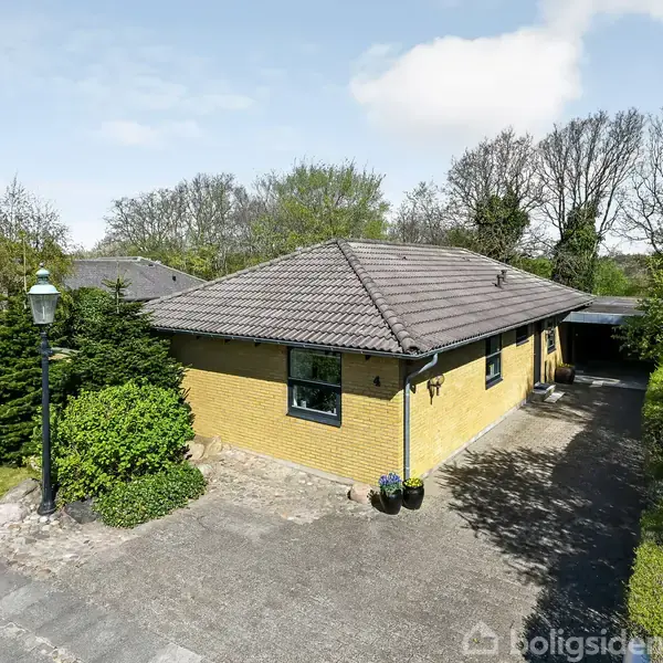 Et gult murstenshus med gråt tag står stille i et grønt, træfyldt område. Beliggende ved en asfalteret indkørsel med en tilknyttet carport. Et skilt er placeret ved siden af.