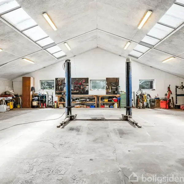 Løfteplatform står tom i en rummelig garage med højt til loftet. Værktøjsbænke og hylder fyldt med værktøj og diverse genstande omkranser væggene. Loftet har ovenlys og belysning.