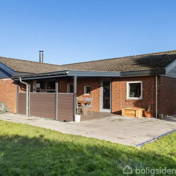 Et rødt murstenshus med skråtag står på en terrasse omgivet af græsplæne. Huset har en skorsten og en overdækket veranda med havemøbler. Klar blå himmel som baggrund.