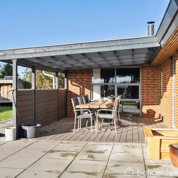 Terrassebord med stole står under en overdækket, trægårdsplads ved siden af et murstenshus. I baggrunden ses en græsplæne og flere huse i nabolaget.