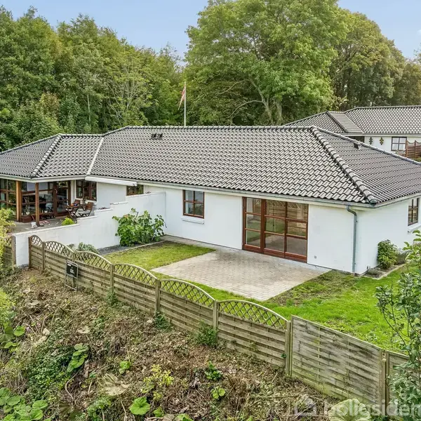Et hvidt hus med sort tag ligger i en frodig have omgivet af træer. En lille terrasse ses ved siden af en græsplæne omgivet af et hegn.