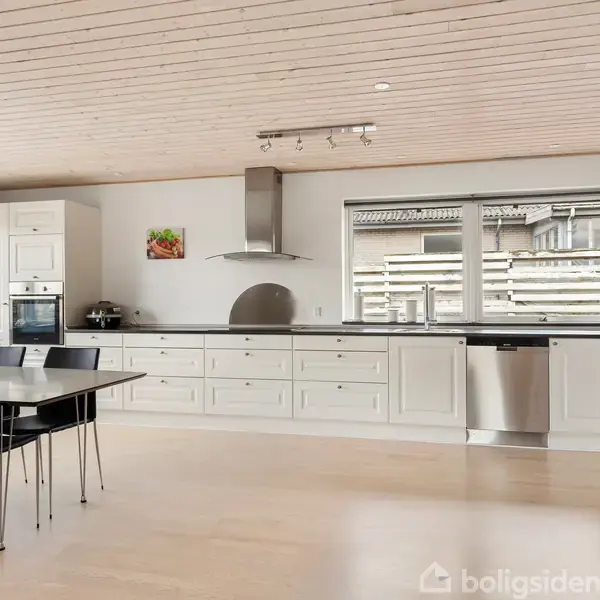 Køkken i moderne stil har et spisebord med sorte stole. Rummet har hvid indretning, rustfrit køleskab, ovn og opvaskemaskine, samt stort vindue, der slipper naturligt lys ind.