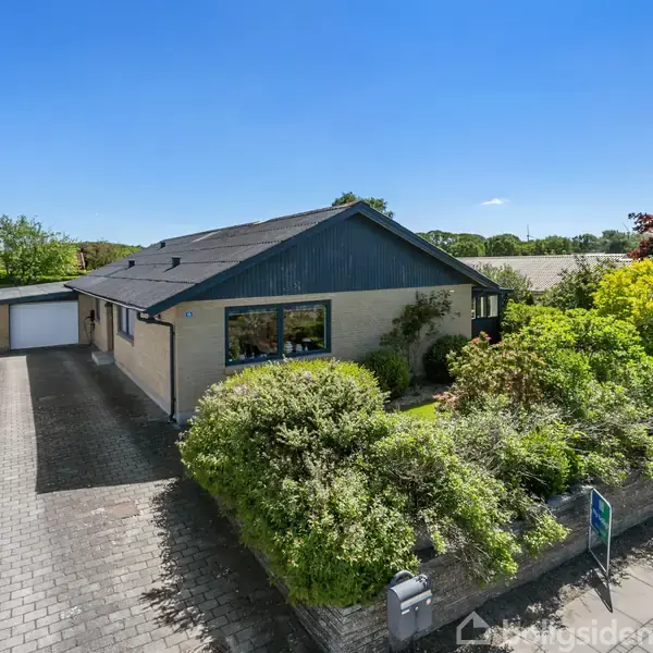 Hus omkranset af frodig have, rummende stor forhave. Indkørsel af fliser foran fører til dobbelgarage. Solrigt vejr med blå himmel.