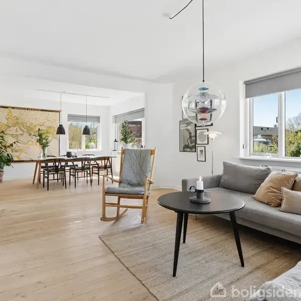 Stue med grå sofa og sofabord med dekorative genstande. En lænestol står ved siden af vindue. I baggrunden ses et spisebord med stole. Store vinduer giver udsigt til haven.