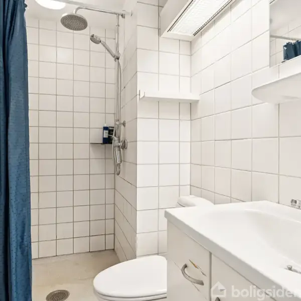 Bruser hænger i brusekabine med hvidt flisebeklædt væg. Toilet og vask beskæftiger rummet med hvide møbler og blå forhæng, skaber et rent, moderne udseende. Ingen tekst.
