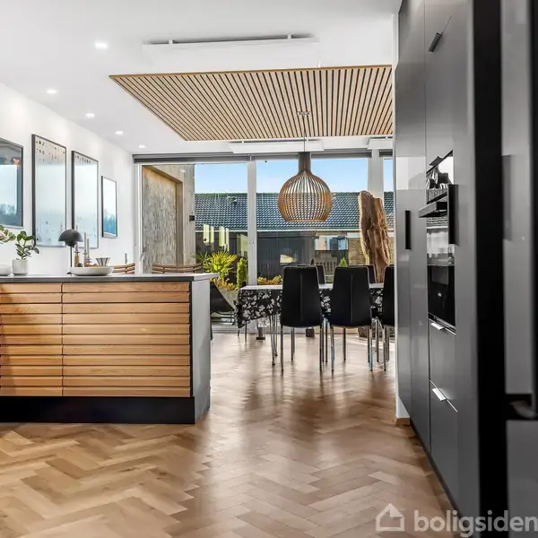 Køkken og spisestue med trægulv, moderne træpaneler på køkkenøen. Sort spisebord med sorte stole står ved store vinduer, der viser udsigt til et hus udenfor.