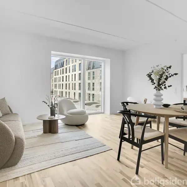 En lys stue med en beige sofa og et rundt sofabord i forgrunden. Et spisebord med stole står ved siden af. Store vinduer viser moderne bygning udefra. Ingen tekst.