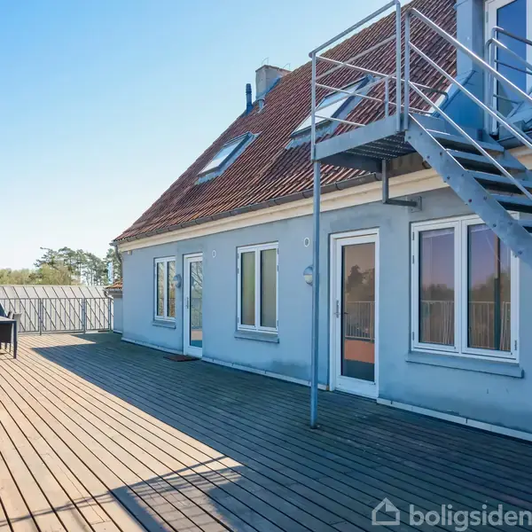 To-etagers hus med udendørs trappe på solrig, træbelagt terrasse. Flere vinduer og døre fører ud til terrassen. Et bord og stole ses til venstre, omgivet af træer.