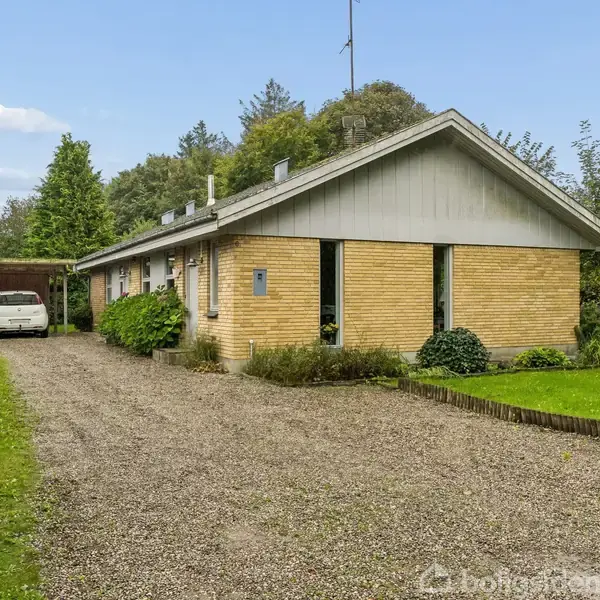 En étplanshus af mursten står stille med bil i carport til venstre. Huset er omgivet af grønne træer og buske med en grusvej foran.