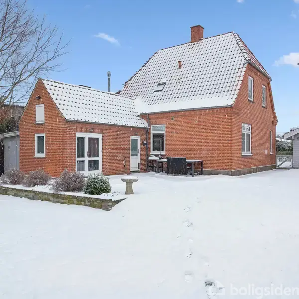Rødt murstenshus med sneklædt tag og have. Huset står stille i en vinterhave med en lille bygning ved siden af. Himlen er klar med spredte skyer.