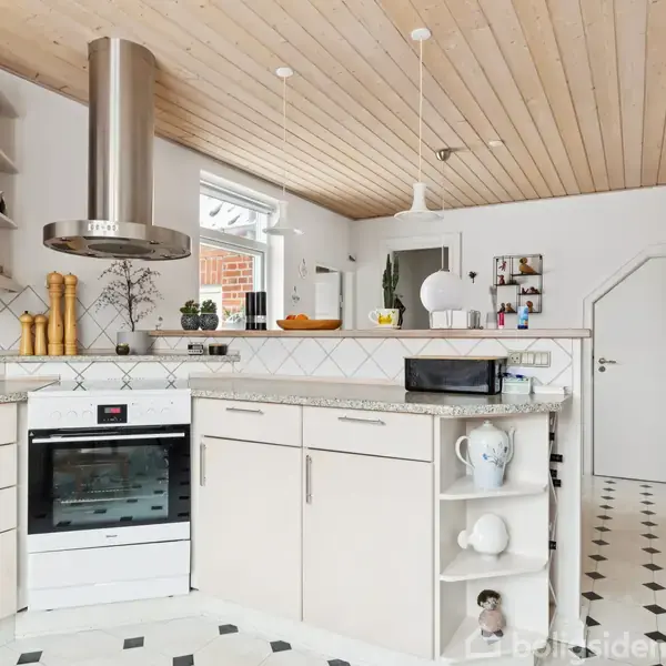 Moderne køkken med hvide skabe og sort-hvidt ternet gulv. Køkkenø med indbygget ovn og emhætte. Grene i vase og salt- og peberkværne på bordet. Lys træloft.