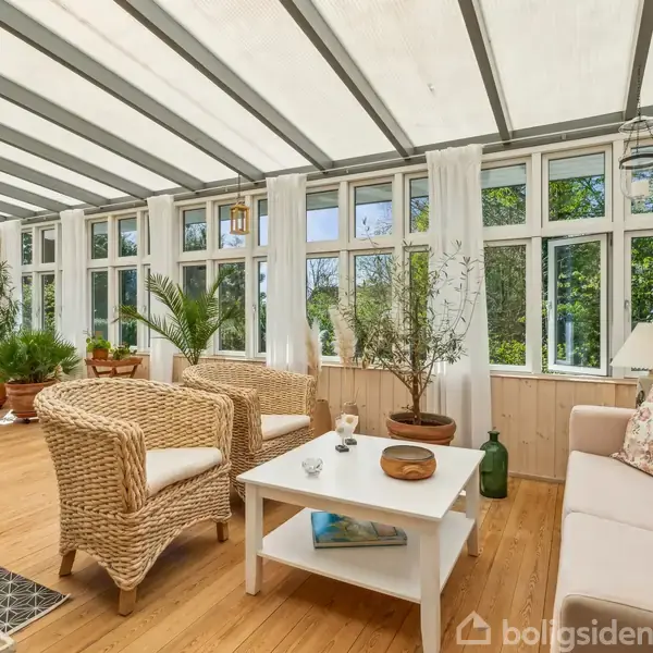 En lys havestue med store vinduer og glasloft rummer kurvestole, en hvid sofa og et sofabord. Grønne planter står rundt omkring, og et spisebord er i baggrunden.