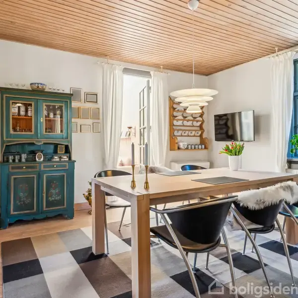 Et spisebord med sorte stole står centralt i et rum med trægulv og loft. En grøn kommode og rektangulært vindue udgør baggrunden. En moderne lampe hænger over bordet.