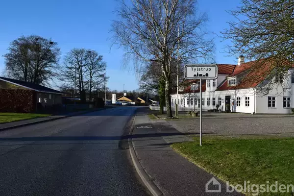 Et vejskilt med teksten "Tylstrup" er placeret nær en vej. Omkring står der træer og en hvid bygning med rødt tag, sandsynligvis et hus.