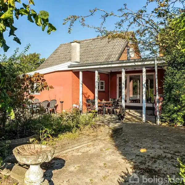 Hus med rød facade og skråtag står midt i en frodig have fyldt med blomster og træer. Foran huset er en terrasse med havemøbler, hvor sollys skinner igennem.